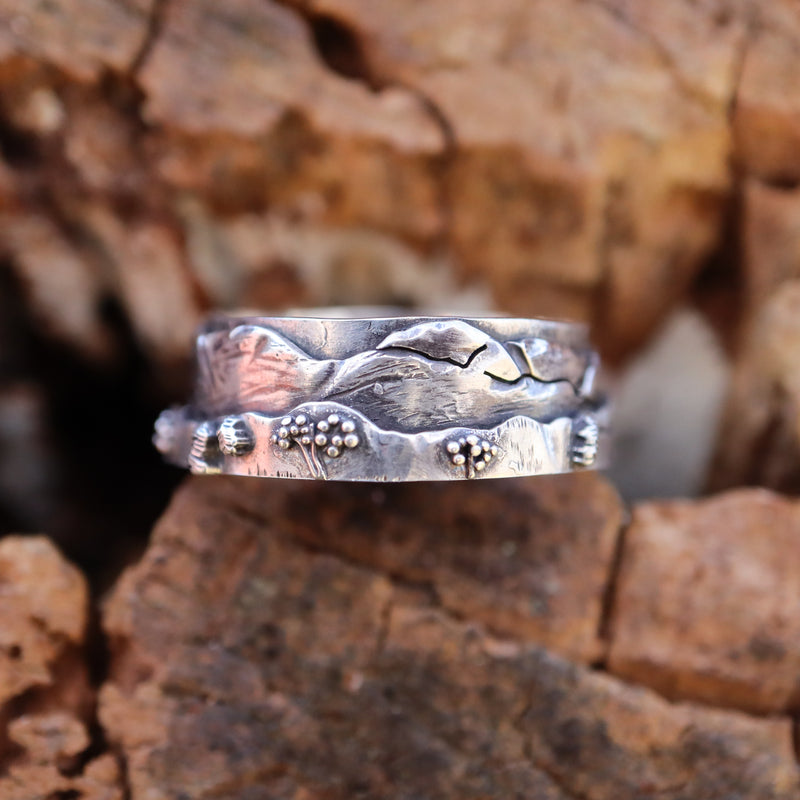 A sterling silver White Mountain range ring with tiny bushes, mountains, and a skyline The ring is shown on a piece of brown wood. 