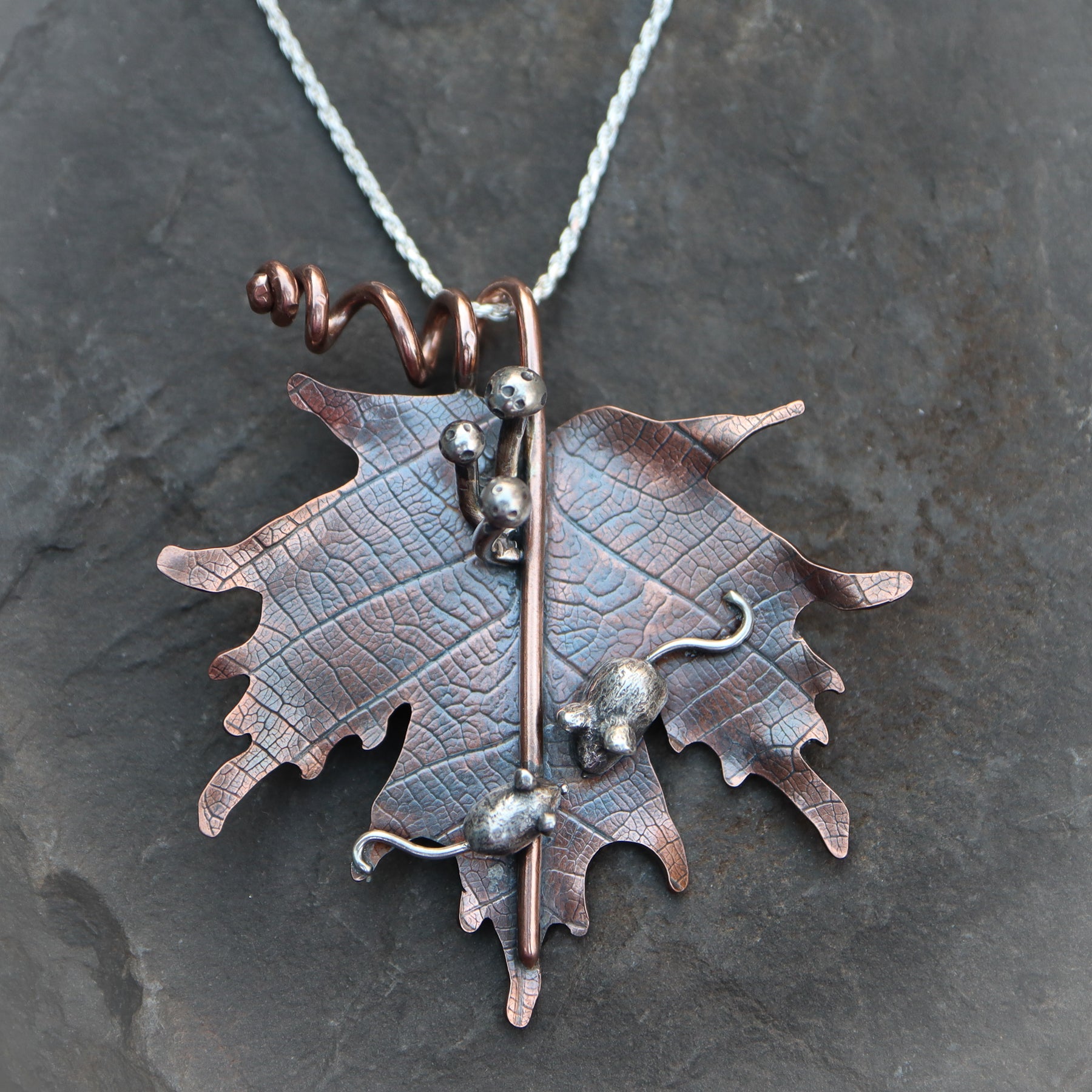A copper maple leaf that is about 2 inches tall and 2 inches wide. On the leaf are two hand carved sterling silver mice, a mother and a baby, snuggled next to each other. Above the mice are three silver mushrooms coming from the stem of the maple leaf. The necklace is shown in a dark grey piece of slate. 