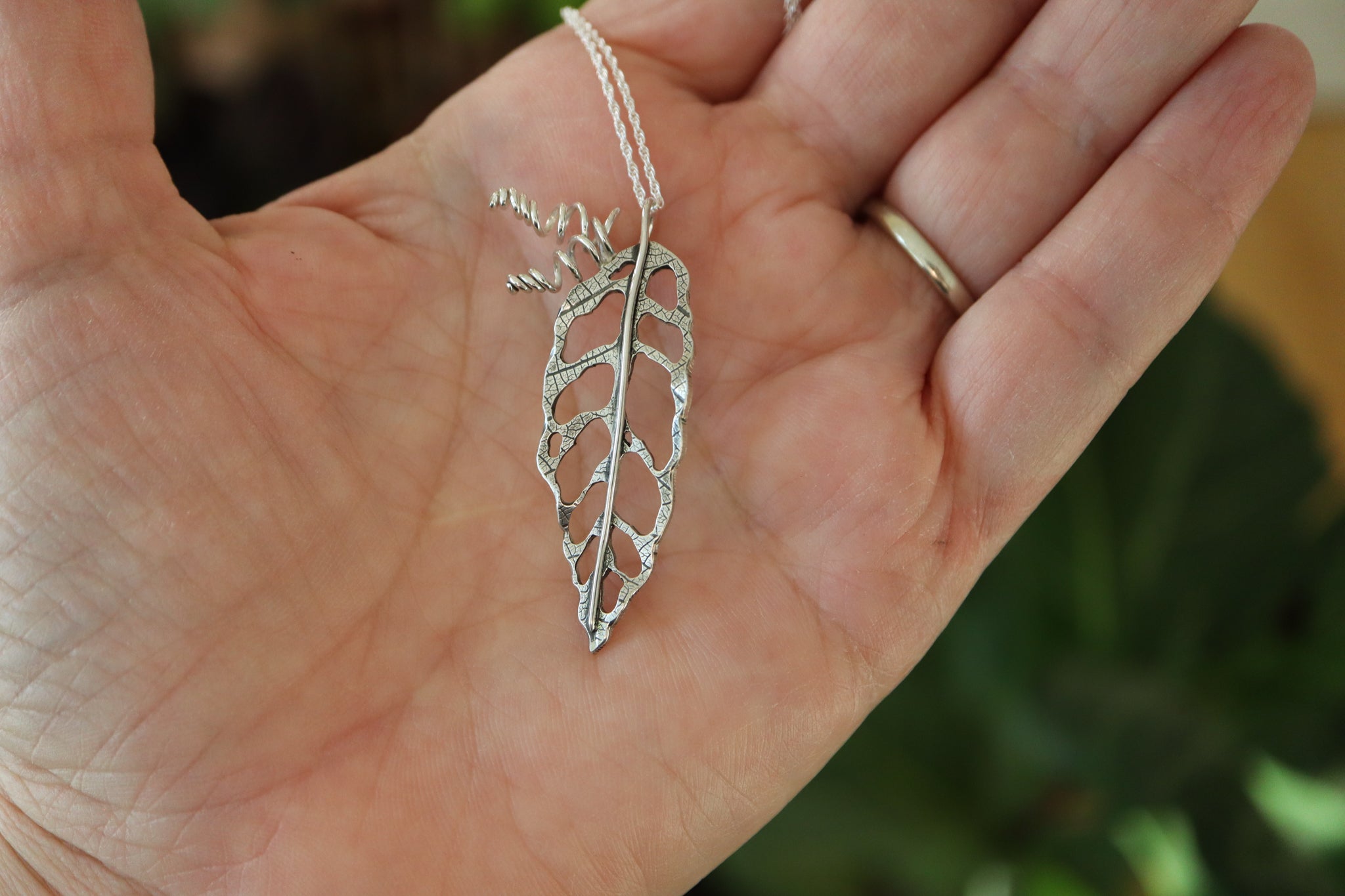 A hand is holding a monstera obliqua sterling silver leaf necklace to show the size. 