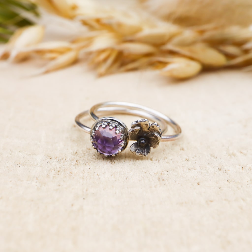 The pansy ring and the purple amethyst ring are shown next to each other. The rings are on a light tank piece of wood. 