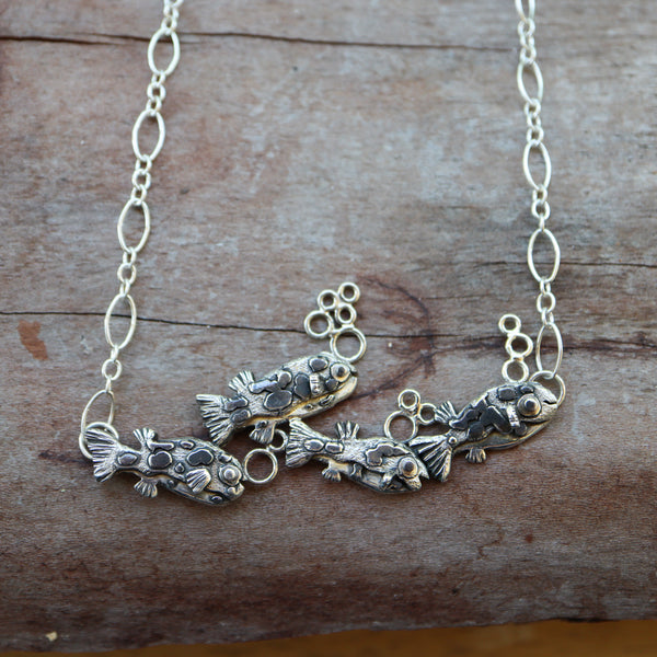 A sterling silver dwarf pea puffer necklace featuring 4 little fish swimming in a row behind each other. All of the fish have a few silver bubbles above them. They are on a silver looped necklace and shown on a piece of dark wood. 