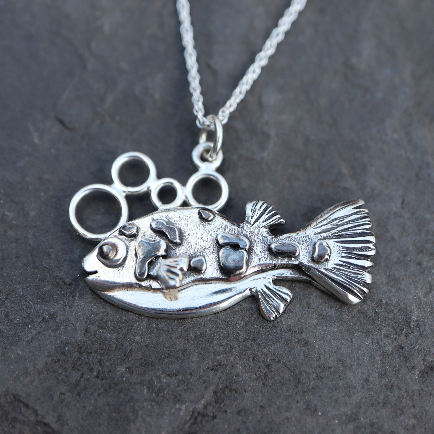 A handmade sterling silver dwarf pea puffer charm necklace is shown on top of a dark slate stone.