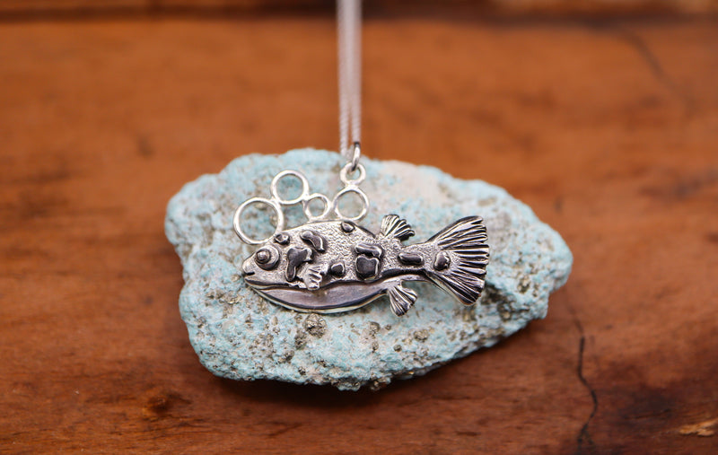 a 1 inch long sterling silver dwarf puffer fish is shown on top of a light blue turquoise stone. 