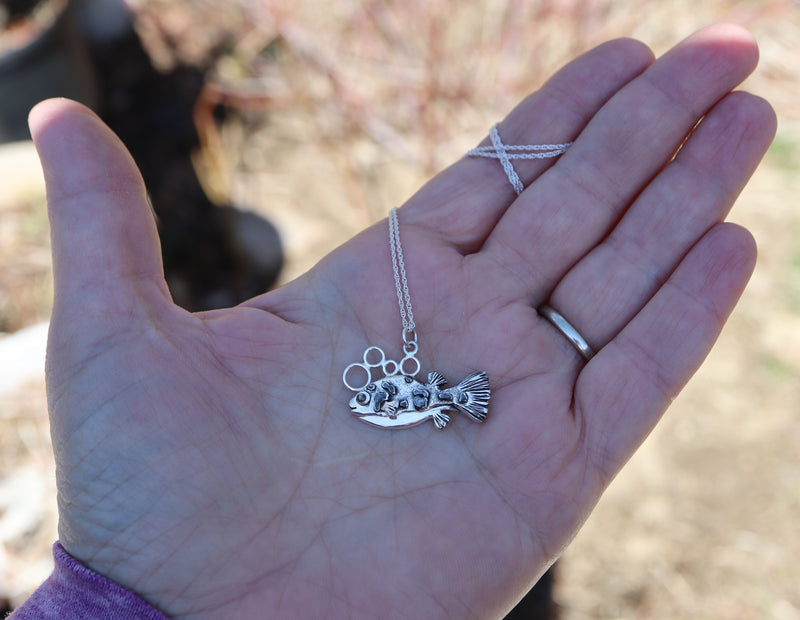 A hand is shown holding a small puffer fish necklace for size reference. 