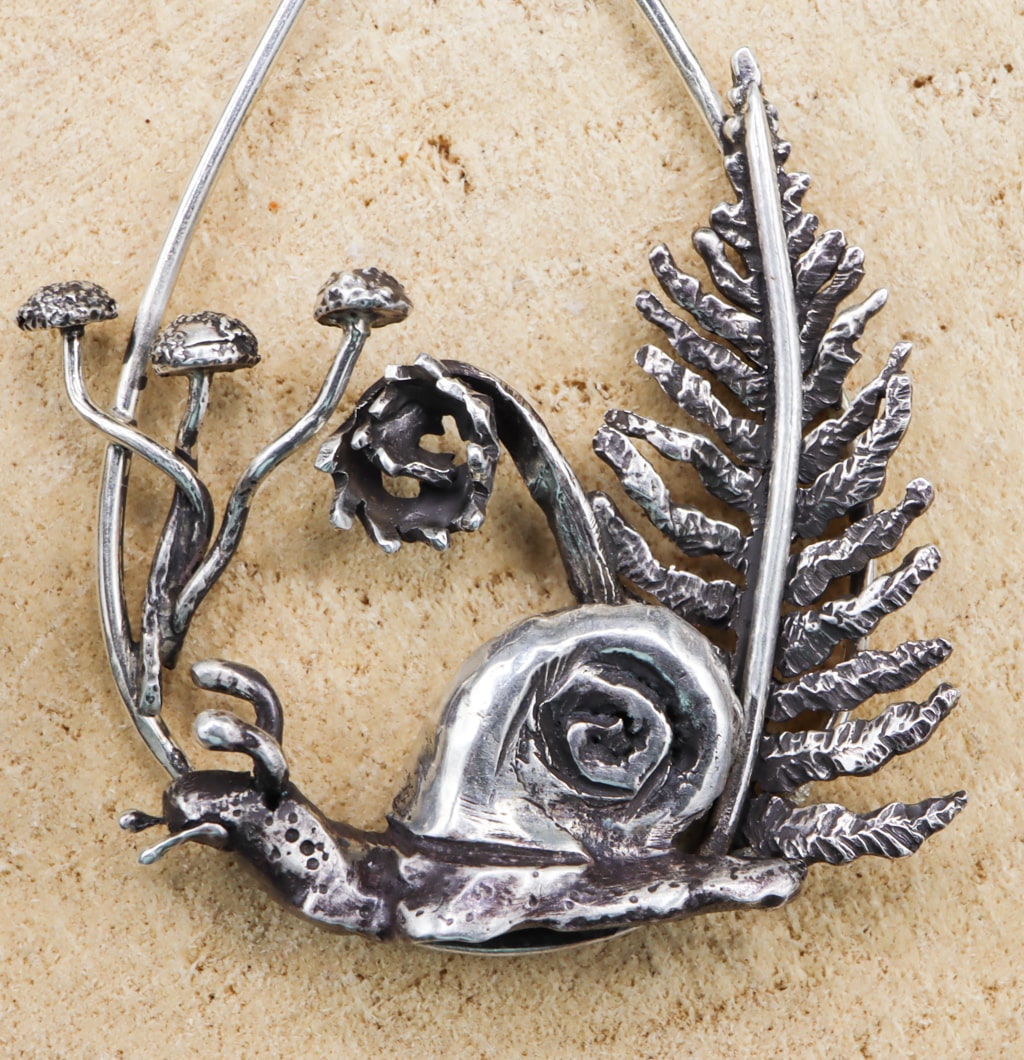 A close up of the sterling silver hand carved snail. Behind him are little mushrooms and ferns. The earring is shown on a light tan piece of wood.