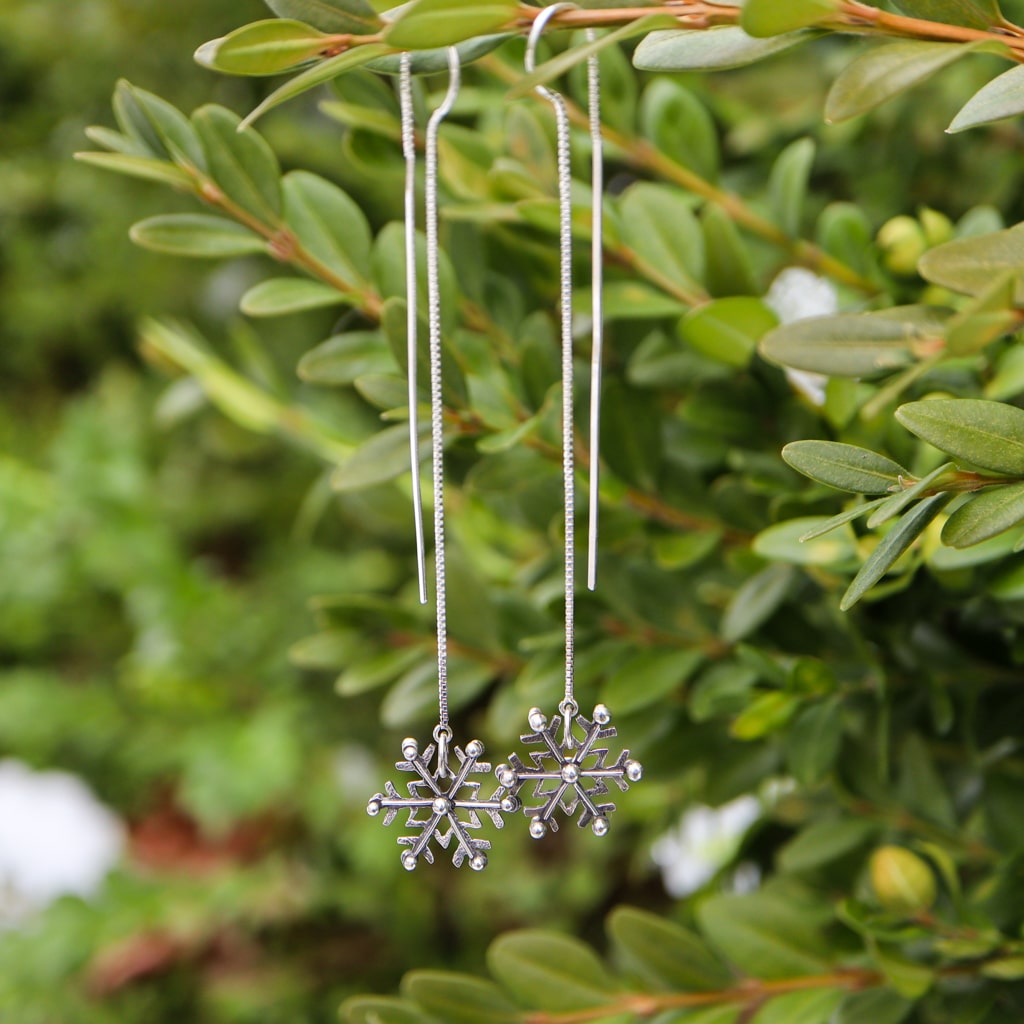 Snowflake ear threader earrings made from sterling silver are about 6 inches long. They are shown hanging on an evergreen bush.