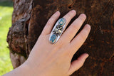 A hand is shown wearing the stag beetle and morenci turquoise with pyrite stone ring. The hand is on a tree trunk .