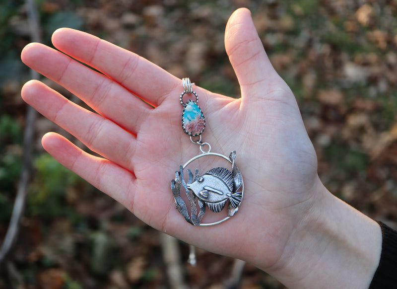 A hand is shown hold the tang fish necklace. It is about 3 inches tall. 