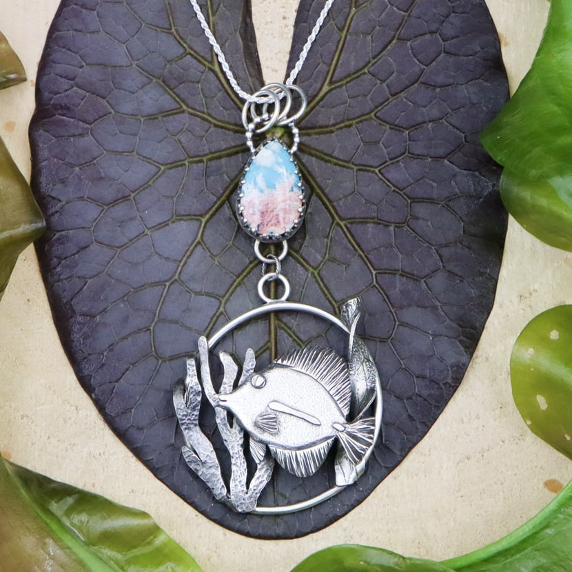 A handmade sterling silver tang fish necklace. The little fish is shown in the center of the pendant and has silver seaweed on each side of her. Above the pendant is a turquoise and quartz doublet stone that looks exactly like the ocean from the sky. The necklace is surrounded by real water plants and on a piece of light tan wood. 