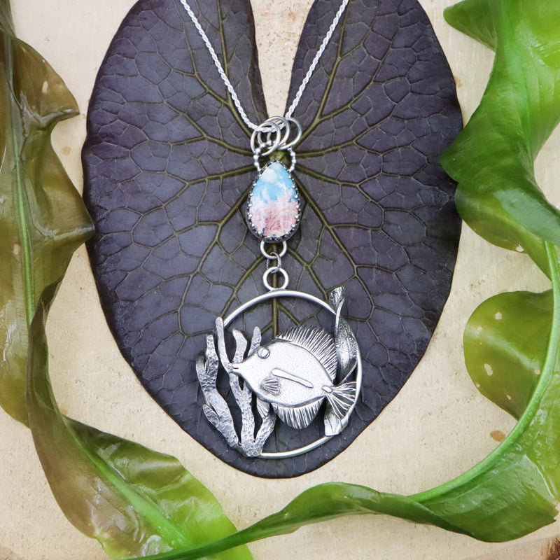A handmade sterling silver tang fish necklace. The little fish is shown in the center of the pendant and has silver seaweed on each side of her. Above the pendant is a turquoise and quartz doublet stone that looks exactly like the ocean from the sky. The necklace is surrounded by real water plants and on a piece of light tan wood. 