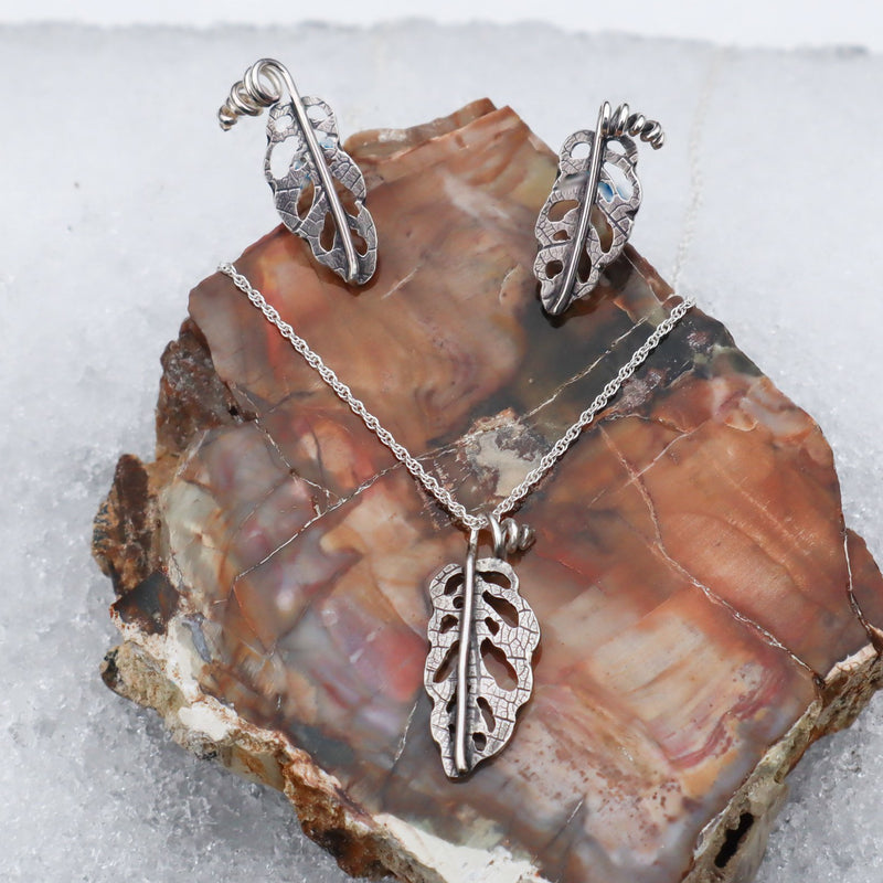 A set of sterling silver jewelry that look like small monstera adansonii leaves. A necklace, earrings, and pendant are shown on a piece of petrified stone in pretty brown colors.