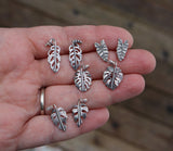 A hand holding 8 pairs of sterling silver plant stud earrings. Monstera deliciosa, adansonii, alocasia polly, and rhaphidophora tetrasperma are shown.
