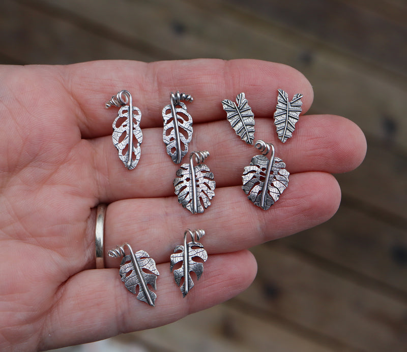 8 pair of earrings are being held in a hand to show the sizes. Monstera adansonii, monstera deliciosa, alocasia polly, and rhaphidophora tetrasperm leaves. 