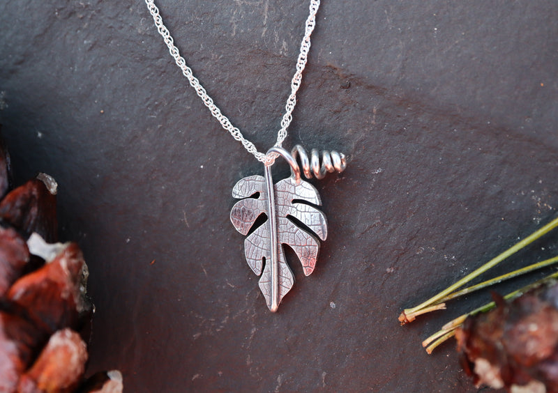 A small sterling silver rhaphidophora tetrasperma pendant with a necklace is shown in front of a dark gray slate tile.