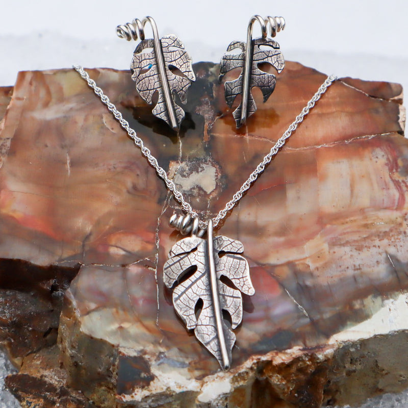 A photo of sterling silver handmade rhaphidophora tetrasperma necklace and earring set. They are shown on a piece of petrified wood with snow in the background.