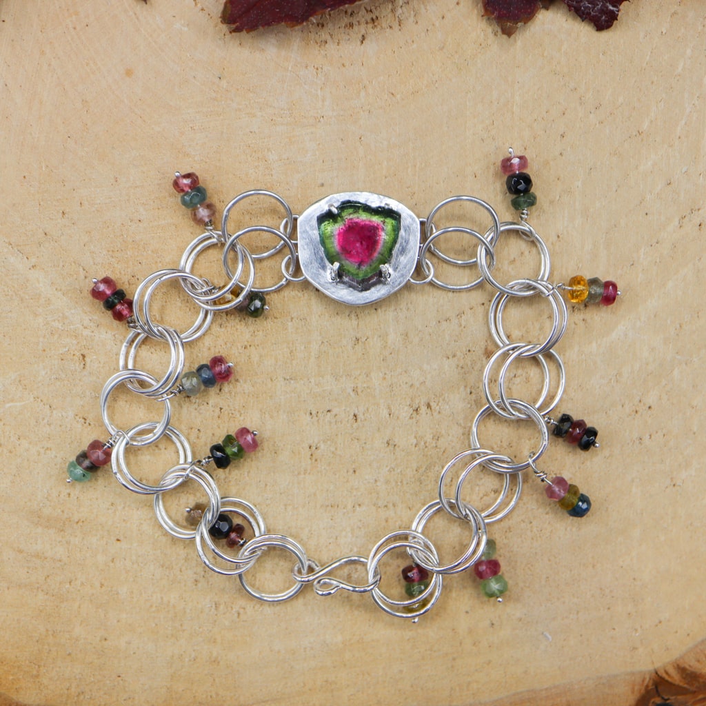 Watermelon tourmaline bracelet with a central pink and green roughly round shape stone. There are large circular links that make up the bracelet. On each link there is a little strand of three multi colored tourmaline stones that move around when the bracelet is being worn. It is shown on a piece of light tan wood. 